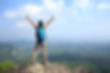 The photo shows a hiker standing triumphantly on a rocky peak with arms raised, overlooking a vast landscape of mountains, valleys, and a distant body of water under a bright blue sky.