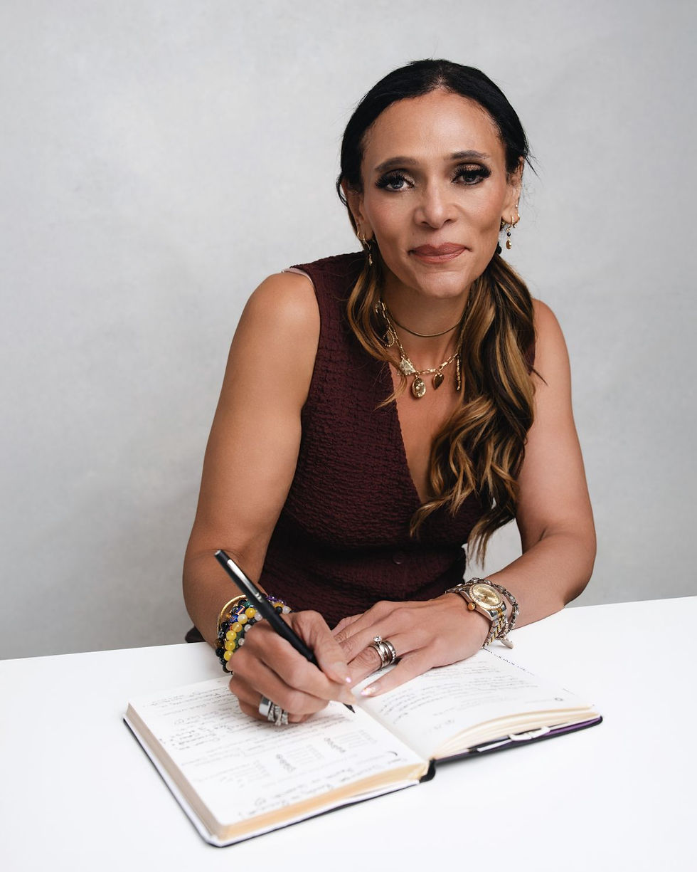 A woman in a sleeveless maroon top sits at a table, writing in an open notebook while smiling slightly at the camera.