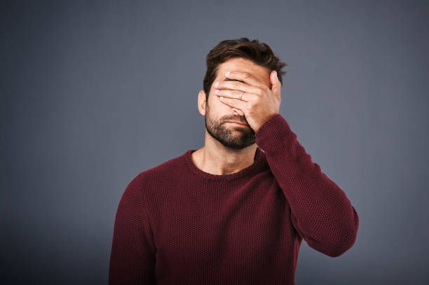 An adult man wearing a maroon long sleeve covering his eyes in in shame.