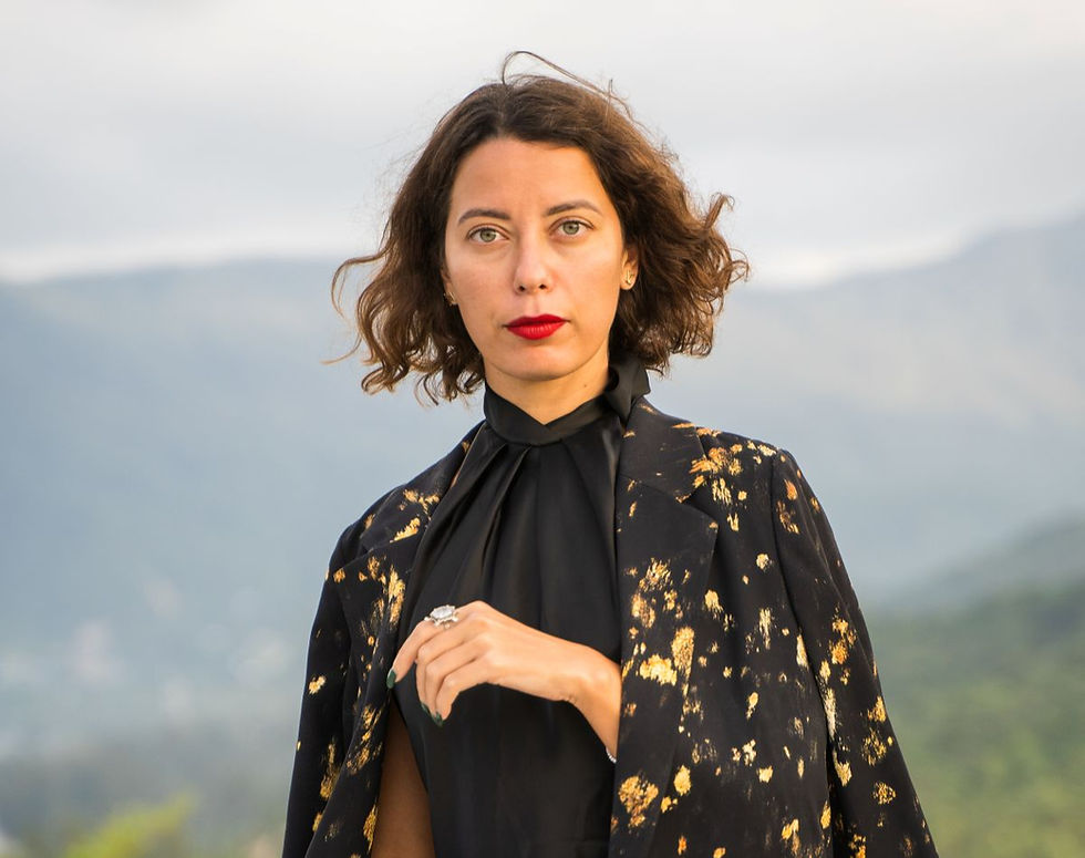 Portrait of a woman with wavy hair in a black-and-gold patterned outfit, standing outdoors against misty mountains.