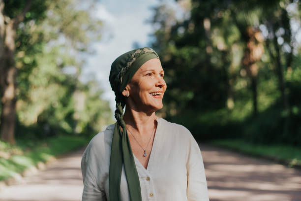 Smiling woman in a green headscarf and white blouse stands on a sunlit tree-lined path outdoors.
