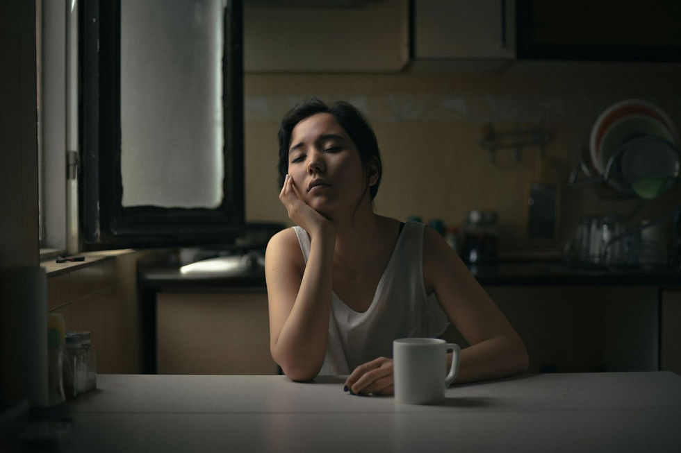 Woman sitting at a kitchen table with a white mug, resting her head on her hand, eyes closed. Natural light through a window creates a calm mood.
