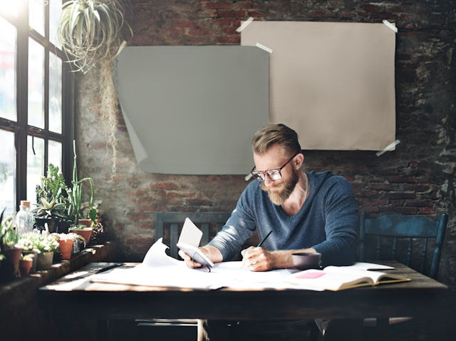 Man with glasses writing at a rustic wooden desk, surrounded by plants and books. Brick wall with posters in the background. Peaceful mood.