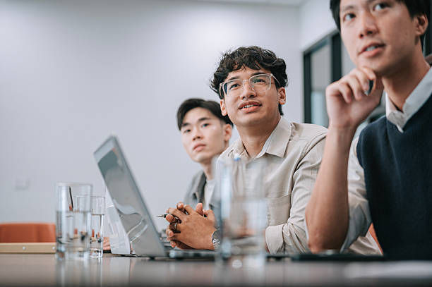 Three young men in a meeting room sit by a laptop and water glasses, one smiling attentively at the table.