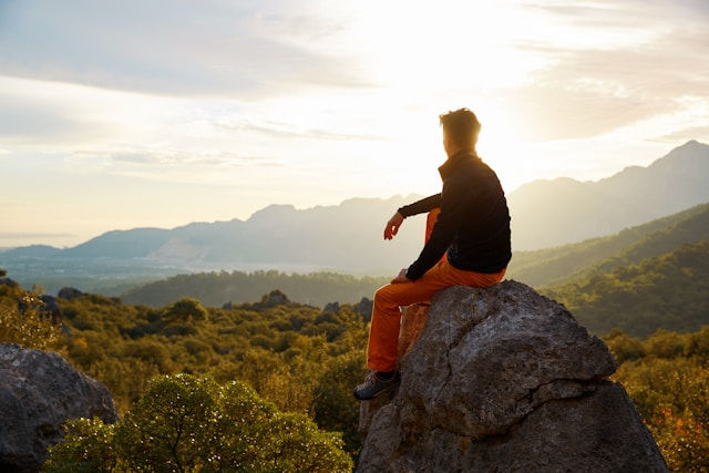 Person in black jacket and orange pants sits on a rock, overlooking a green valley at sunset. Mountains and sun in background, serene mood.