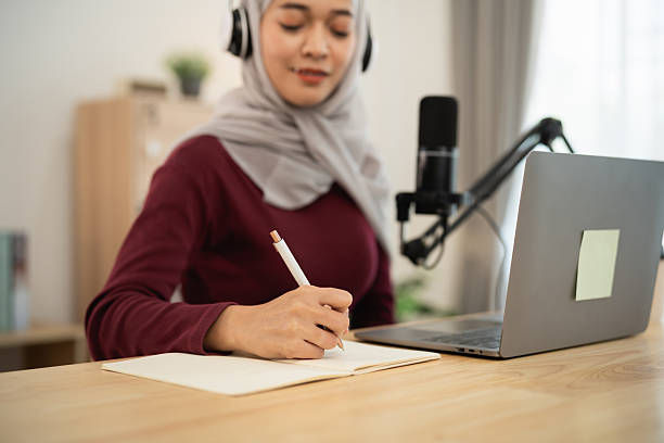 Woman in a maroon top and hijab writing in a notebook, wearing headphones in front of a laptop and microphone, in a cozy room.