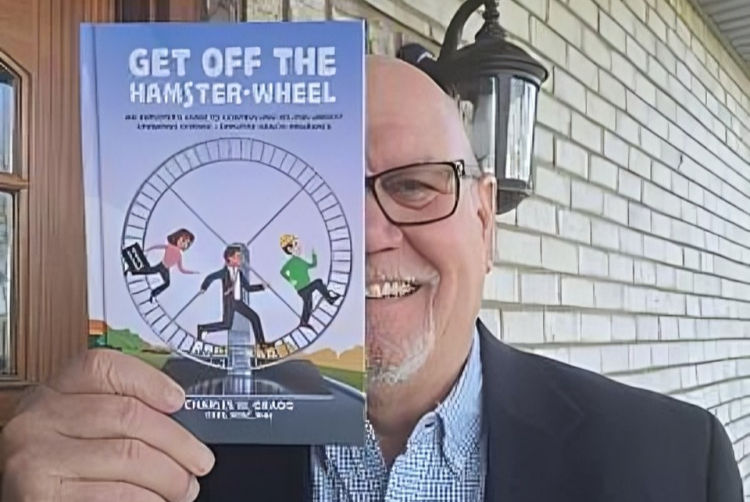 Man smiling, holding a book titled "Get Off the Hamster-Wheel" with a cartoon cover. Background shows a brick wall and outdoor light.