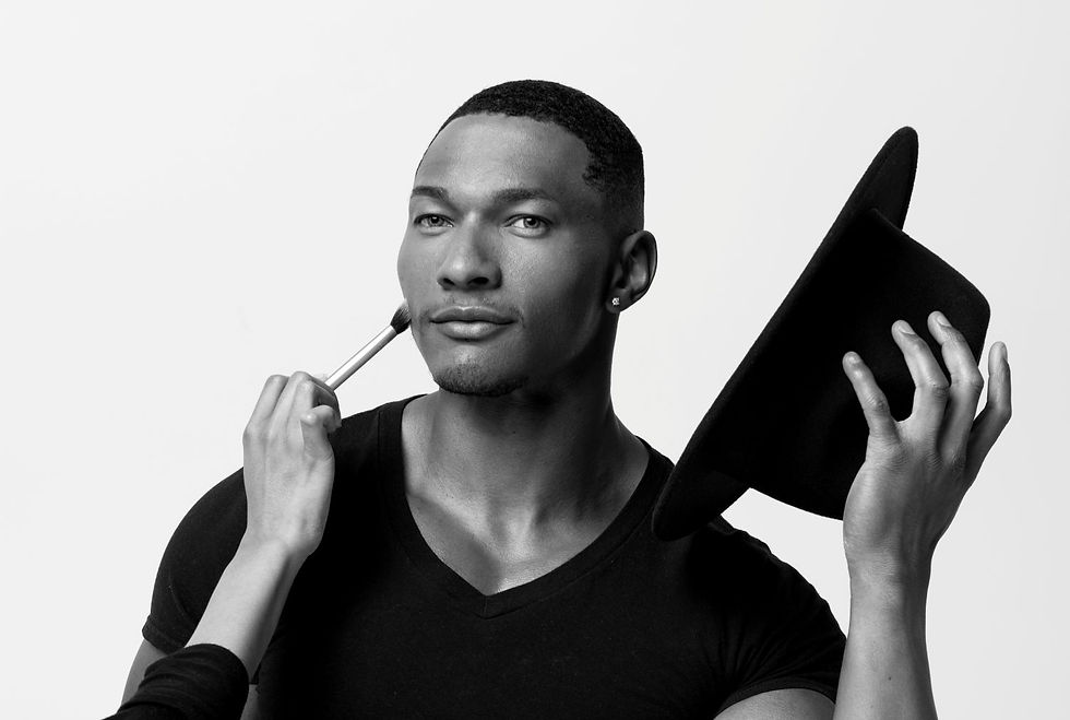 Man in black shirt holds hat, gets makeup applied with brush on face. Monochrome image, serene expression, plain background.