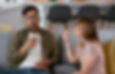 A man and woman on a gray sofa, gesturing as if speaking, with a kitchen in the background. The mood is conversational.