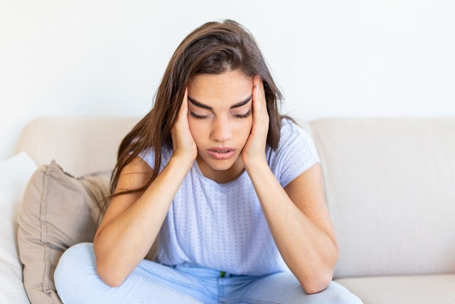 A woman sits on a couch with her head in her hands, appearing stressed or upset.