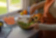 Woman in an orange apron mixes salad in a glass bowl by a sunny kitchen window, with carrots and roasted chicken nearby.