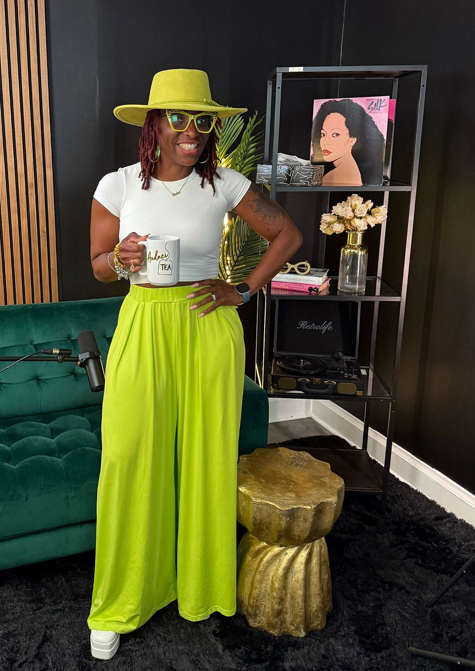 Woman in lime outfit and hat holds "AudaciTEA" mug, smiling in a chic room with art, books, and a microphone. Vibrant, cheerful mood.