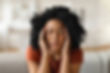 A woman with curly hair is sitting with her eyes closed, pressing her temples with both hands, appearing to be in pain or stress.
