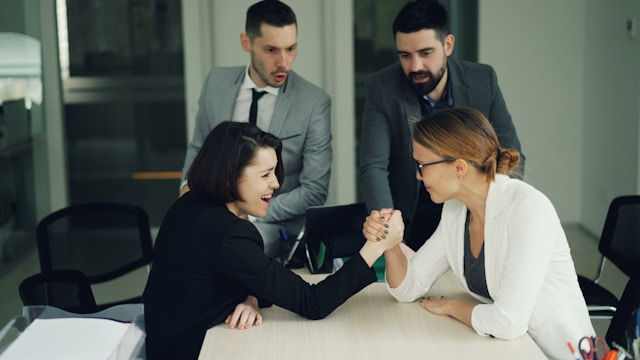 Two women arm wrestle at a conference table while two men watch in an office, the mood tense and competitive.