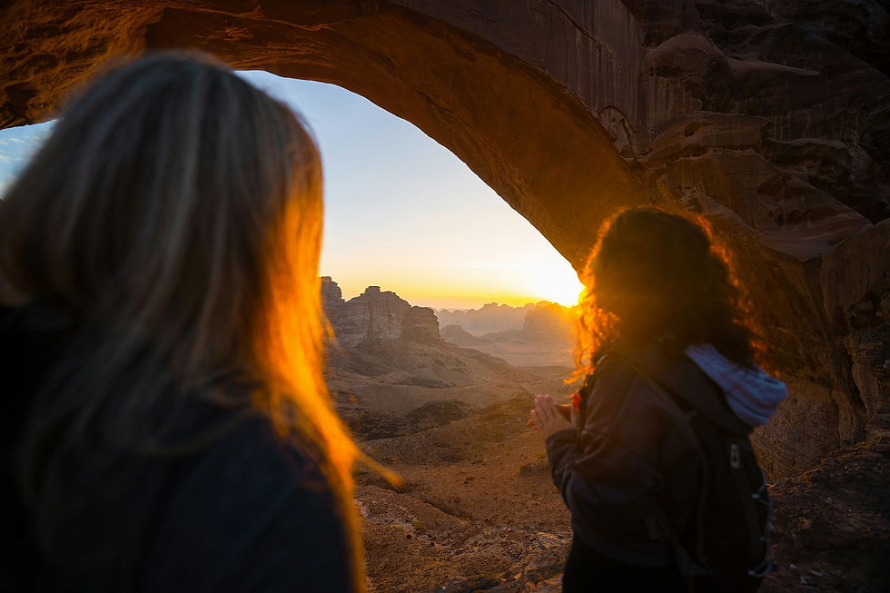 Two people stand under a rock arch, gazing at a sunset over a desert landscape. Warm, golden light highlights their silhouettes.