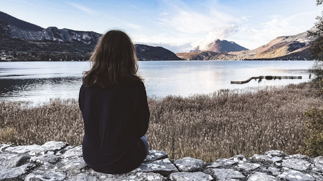 Person sitting on a stone wall, facing a serene lake surrounded by mountains. Overcast sky; mood is calm and contemplative.