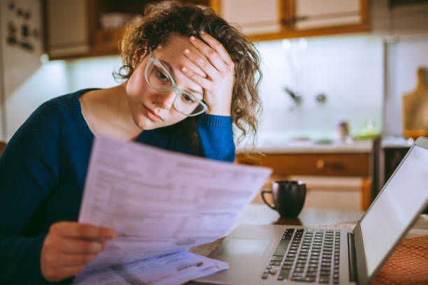 Woman in glasses looking stressed, holding papers in a kitchen with a laptop and mug nearby. Background has warm lighting and wooden cabinets.