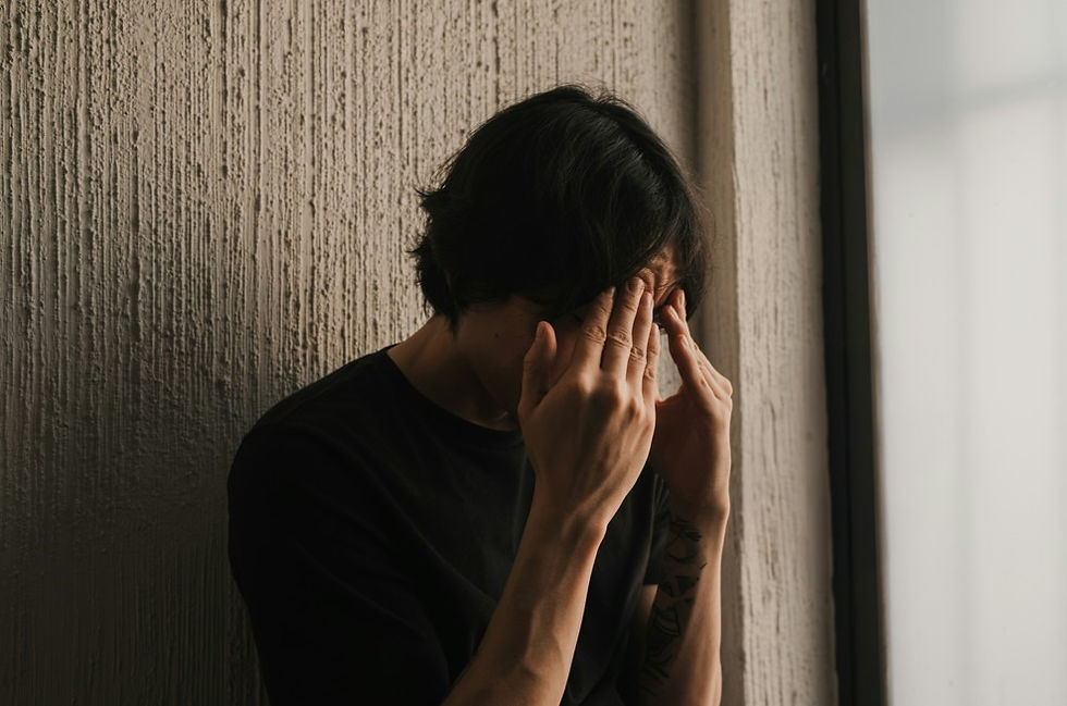 Person in a black shirt covers their face beside a textured wall and bright window, suggesting stress or sadness.