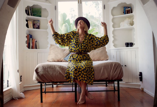 A woman in a floral dress and hat stretches joyfully while sitting on a bed in a cozy, sunlit room.