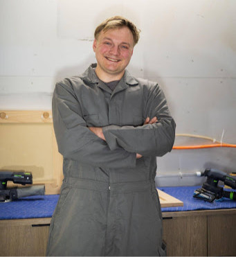 Smiling man in a gray jumpsuit stands with arms crossed in a workshop. Tools and wood pieces are on the bench behind him.