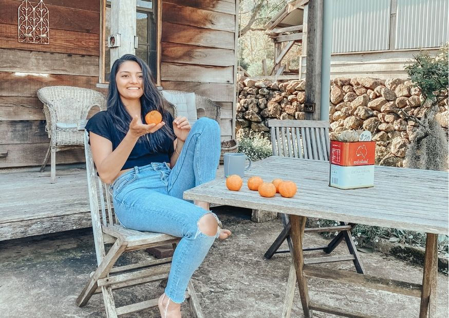 Woman sitting on wooden chair, holding an orange, surrounded by orange fruits on table. Rustic setting with wood cabin and stone wall. Casual mood.