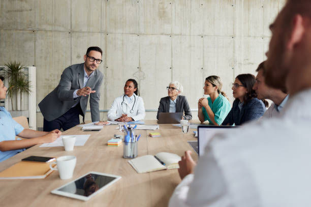 Man in gray blazer presents to a diverse team seated around a conference table in a modern office with a concrete wall.