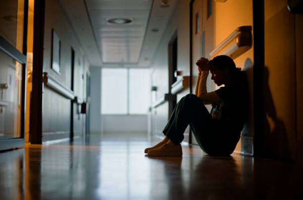 Overwhelmed woman sitting at a laptop with her hands on her head while coworkers move quickly around her in a busy office.