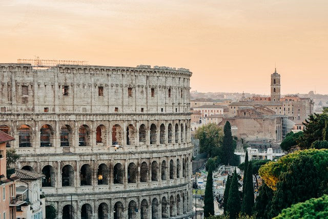Roman Colosseum at sunset beside Rome’s skyline, with trees and a warm orange sky.