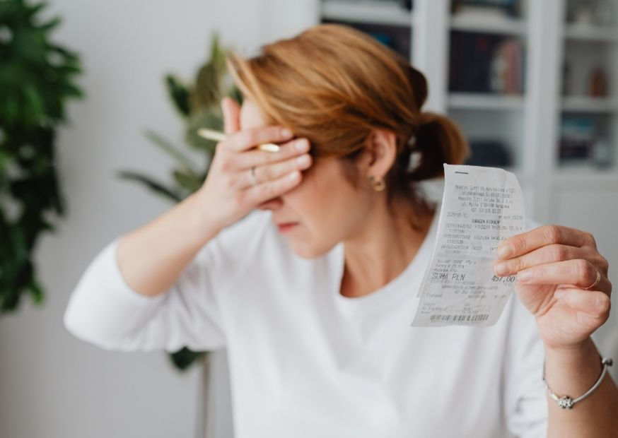 Woman holds receipt, covering face with hand, appears stressed. Dressed in white, indoor setting with plants and shelves in background.