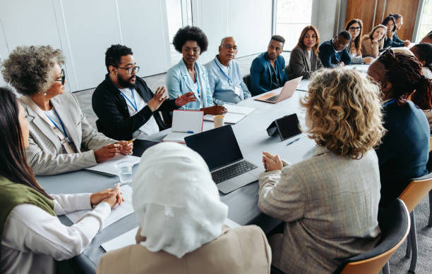 Diverse group in a business meeting around a long table. Laptops open, one person is speaking. Bright room with natural light, professional mood.