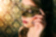 Woman wearing an ornate gold masquerade mask touches her face. Background features an intricate golden pattern. Elegant and mysterious mood.