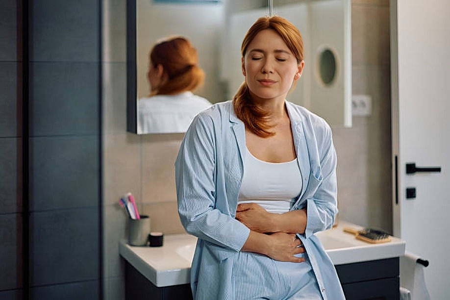 Woman in bathroom holding her stomach, eyes closed in discomfort. Wearing blue pajamas, standing near a sink with toothbrushes. Calm setting.