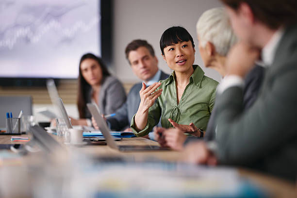 People in a meeting room are discussing at a table with laptops. One person in a green shirt is speaking, creating a collaborative mood.