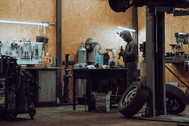 Mechanic in a dim workshop inspects a tool, surrounded by equipment and spare tires. Wooden walls and scattered tools deepen the industrial vibe.