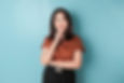 Young woman in a brown top and black pants poses thoughtfully against a light blue background, hand on chin and smiling subtly