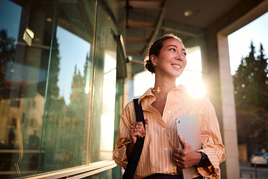 Woman in a striped shirt smiles while holding a tablet, walking in sunlight through an urban setting with glass reflections.