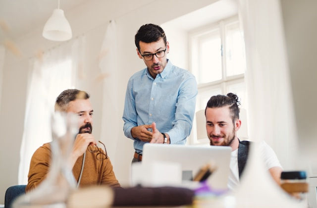 Three men in an office discuss work at a computer. One stands gesturing, two sit focused. Bright room, white walls, relaxed mood.