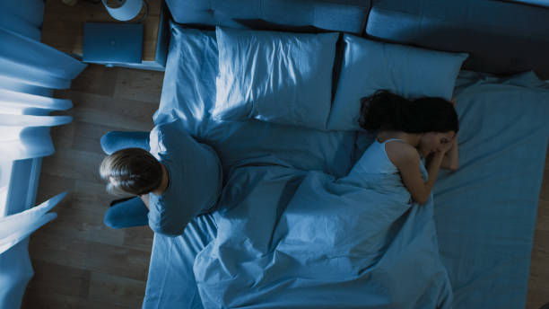 A couple is in bed. One person sits awake, looking away, while the other faces the pillow, asleep. The scene is dimly lit in blue tones.