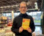 Man smiling and holding a book titled "You Are Not Depressed, You Are Un-Finished" in a bookstore. Shelves and large windows visible.