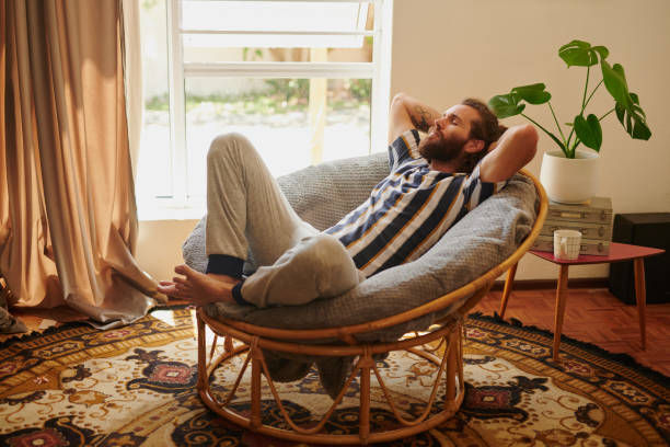 Man relaxing in a round wicker chair in a cozy room, wearing striped shirt and gray pants. Sunlight streams through a window; plant nearby.