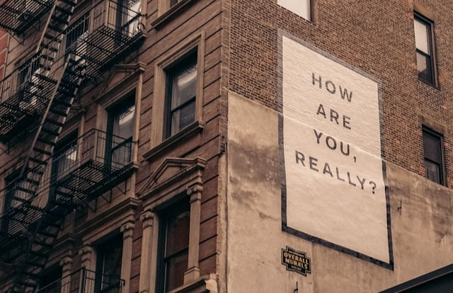 A large mural on a brick building asks, "How are you, really?" A fire escape is visible on the adjacent building.