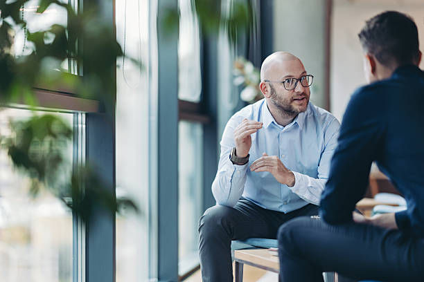 Two men in an office by large windows talk seriously, one gesturing while seated, with soft natural light and blurred plants.