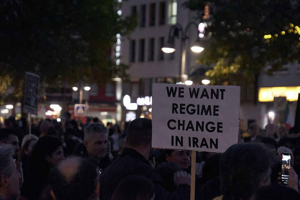 Protesters in a city hold a sign reading "We want regime change in Iran" at night. Dimly lit streetlights set a serious mood.