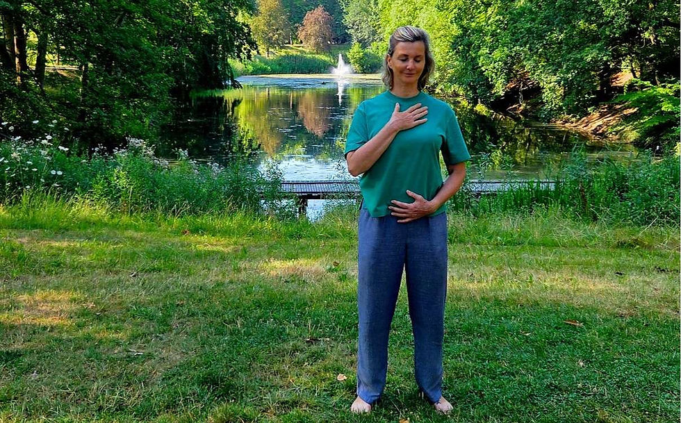 Woman meditates barefoot on grass by a pond fountain in a lush green park, hand on chest and abdomen, calm expression