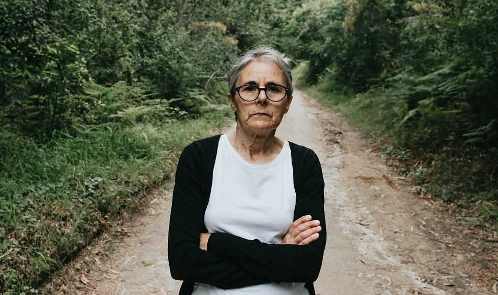 Older woman with glasses stands arms crossed on a dirt forest path, looking serious and concerned.