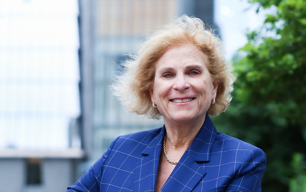 Smiling woman in a blue checkered blazer, outdoors with blurred buildings and greenery in the background. Bright and confident mood.