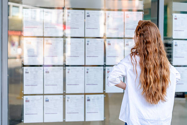 Woman with long hair studies a wall of posted notices on a glass window, likely job listings, in a bright city storefront.