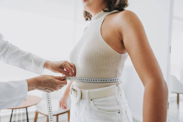 Clinician measures a woman's waist with a tape measure in a bright white clinic; she wears a ribbed sleeveless top and white jeans.