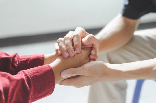 Two people hold hands for comfort in a bright room. One wears a red sleeve, conveying a supportive, caring mood.