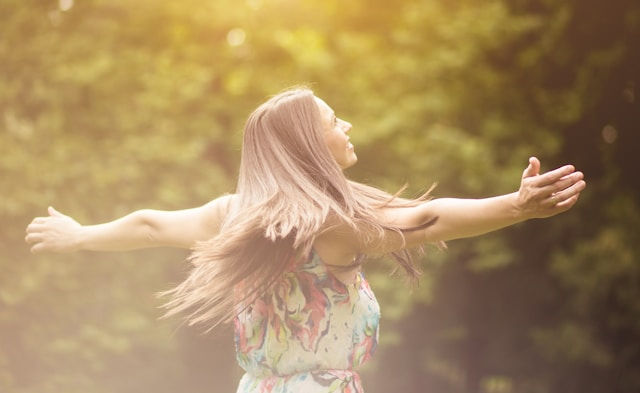 Woman in a colorful dress spins joyfully with arms outstretched in a sunlit, blurry green forest. Bright, serene atmosphere.